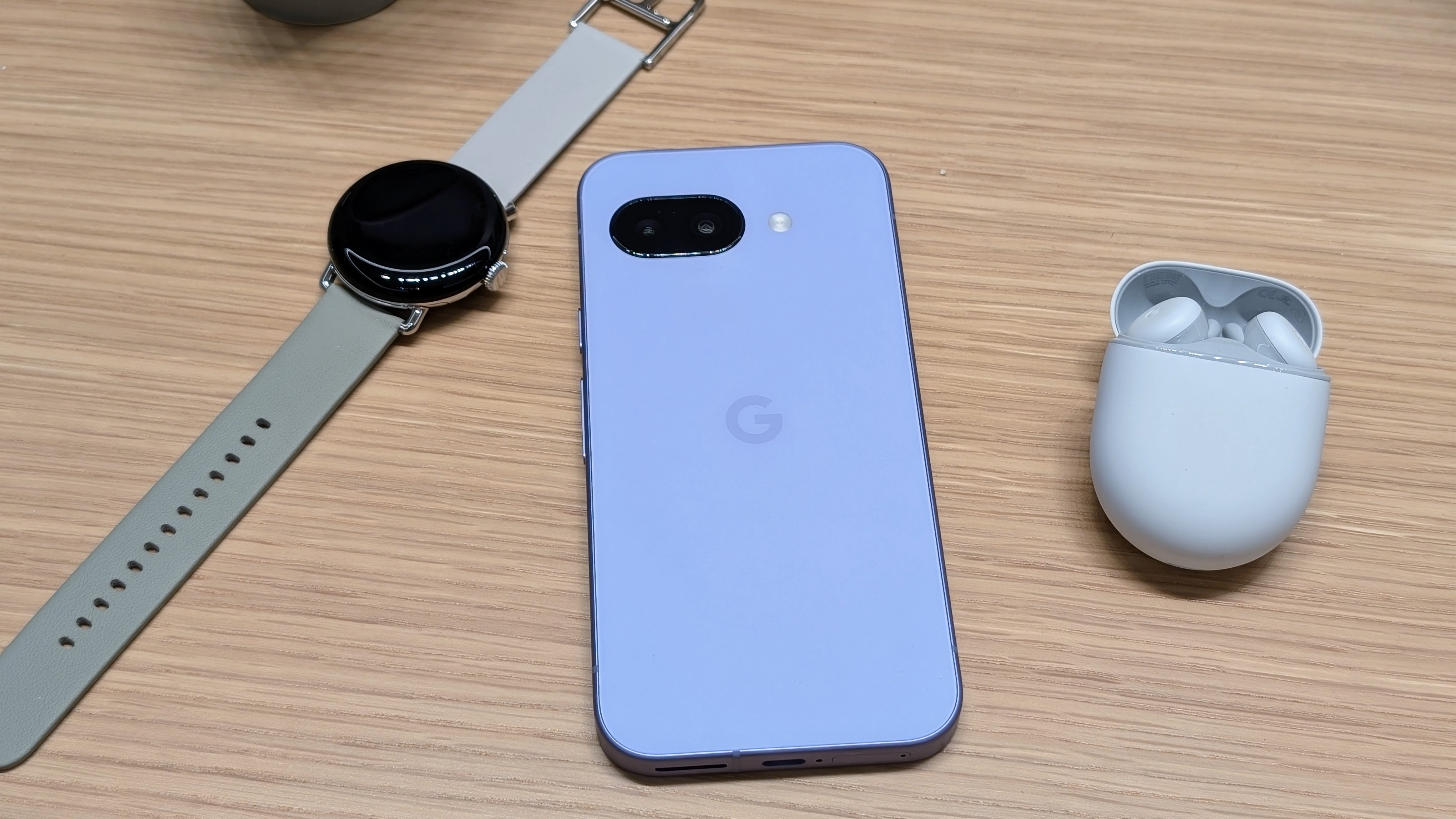 The Google Pixel Watch 3 with gray Active band (left), Iris Google Pixel 9a (center), and Google Pixel Buds (right) sitting close together on a desk. 