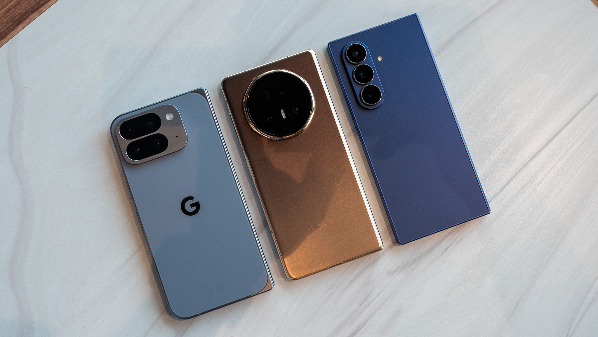 Comparing the size and thickness of the Google Pixel 10 Pro Fold (light blue), Honor Magic V5 (gold/copper), and Samsung Galaxy Z Fold 7 (dark blue)