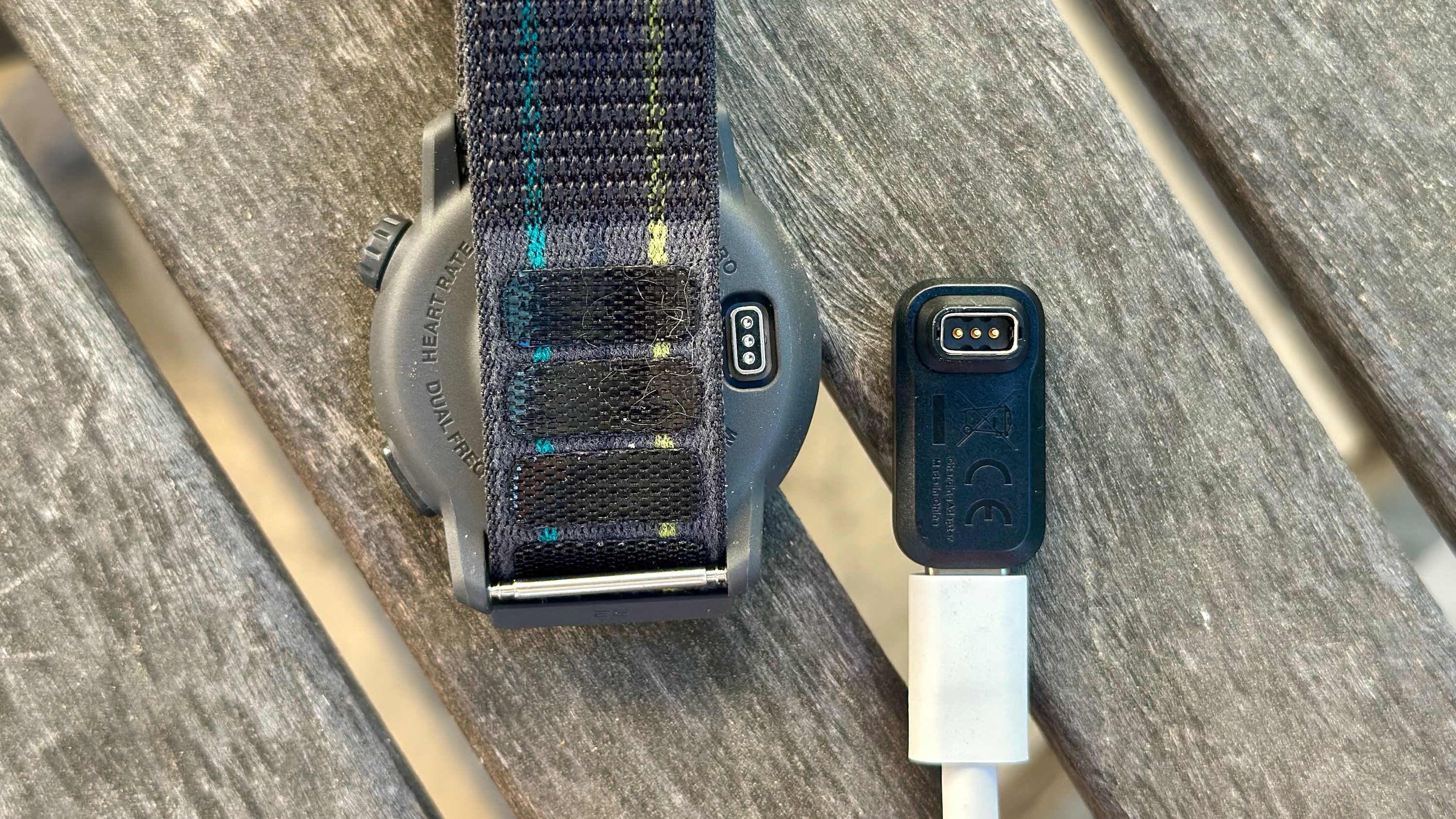 The USB-C to COROS A1 Charging Adapter with a USB-C cable plugged into it, sitting next to an upside-down COROS PACE 3 smartwatch with the proprietary pin charging port visible.